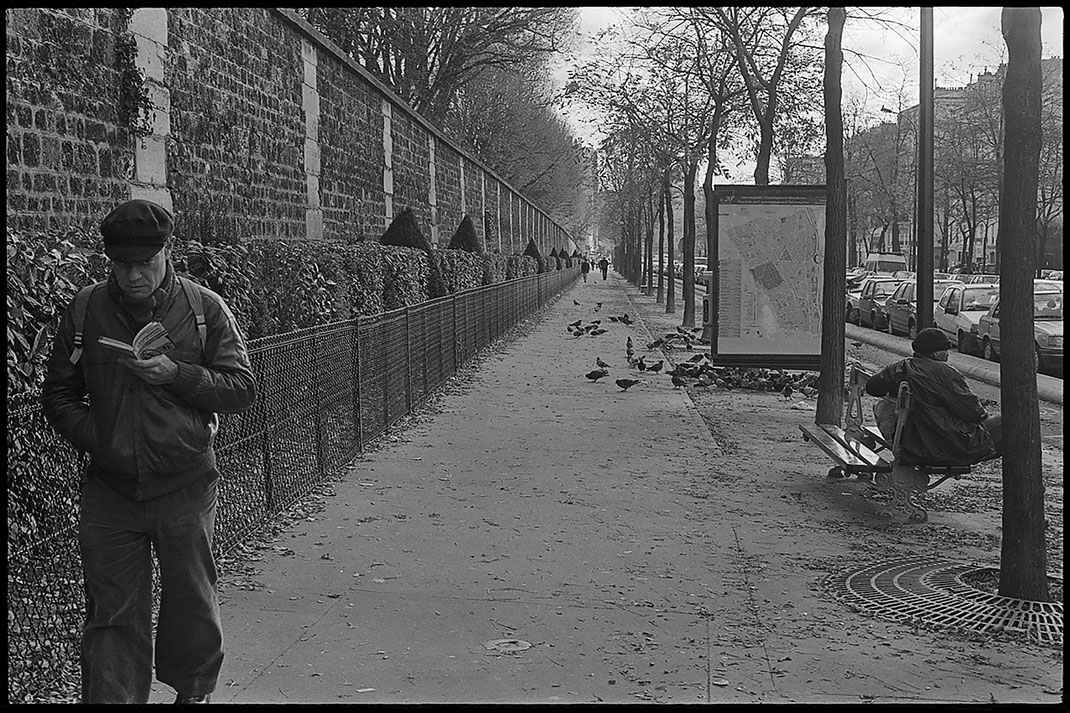 Photographie noir et blanc d'un passant qui lit un livre en marchant sur un trottoir le long du cimetière du Père Lachaise à Paris. On voit quelques dizaines de  pigeons au loin ainsi qu'une personne assise sur un banc à droite de l'image.