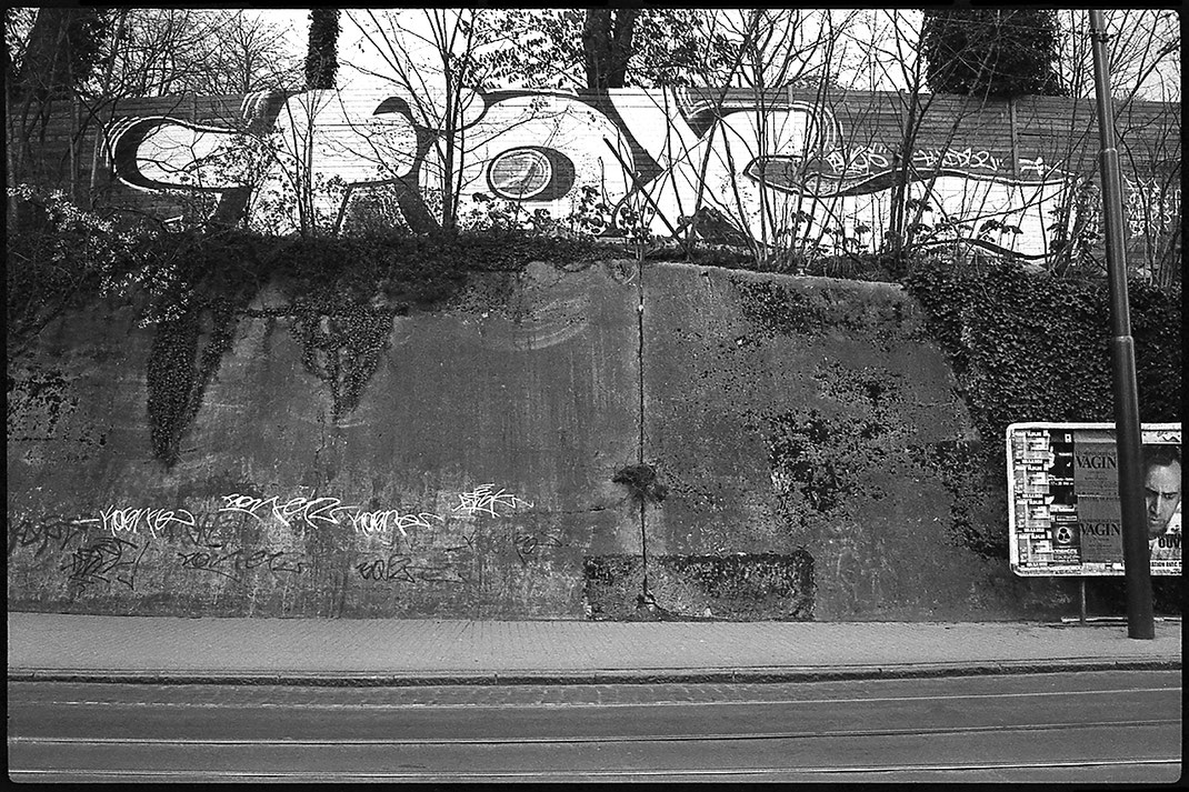 Photographie noir et blanc d'un énorme graphe  "Fox" près du pont de Saint-Job à Uccle, Bruxelles. On voit aussi un panneau de publicités culturelles, avec des affiches "Les monologues du vagin" et "A tombeau ouvert" avec Nicolas Cage.