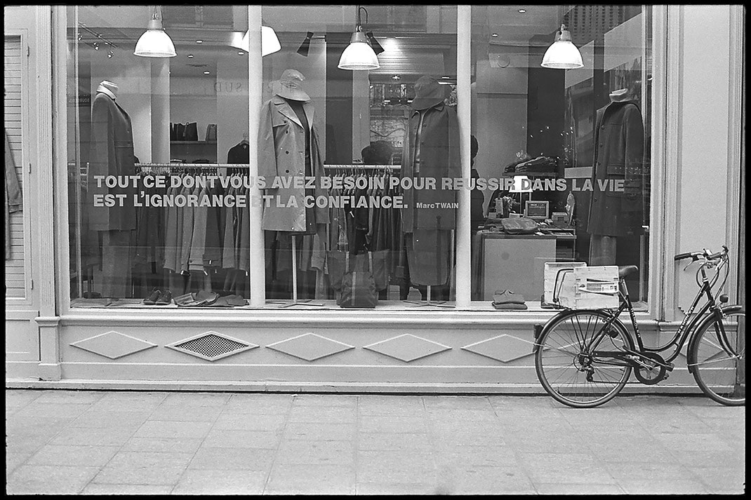 Photographie noir et blanc, vitrine d'une boutique de vêtements sur laquelle est inscrite une citation de Marc Twain " Tout ce dont vous avez besoin pour réussir dans la vie est l'ignorance et la confiance."