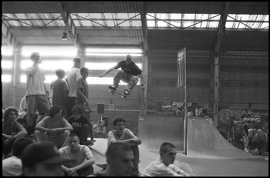 Photographie noir et blanc d'un skateur, effectuant un énorme ollie dans un skatepark lors d'une démo. On voit des spectateurs assis à l'avant-plan qui ne voient pas le skateur en l'air derrière eux. Bruxelles