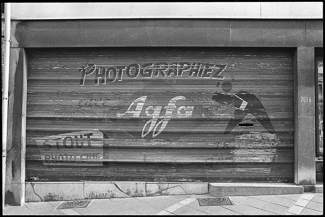 Photographie noir et blanc d'une vieille devanture de magasin de photo. Sur le volet en bois, une peinture sur laquelle on peut lire "Photographiez Agfa", Chaussée de Wavre 261 A, Bruxelles, Belgique.