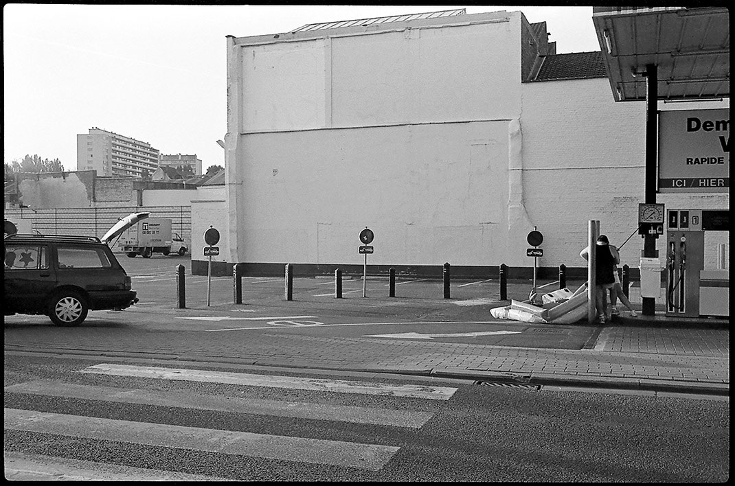Photographie noir et blanc, un couple gonfle une grande piscine gonflable dans une pompe à essence d'un magasin Colruyt. On voit la voiture avec le coffre ouvert sur la gauche de l'image. Bruxelles