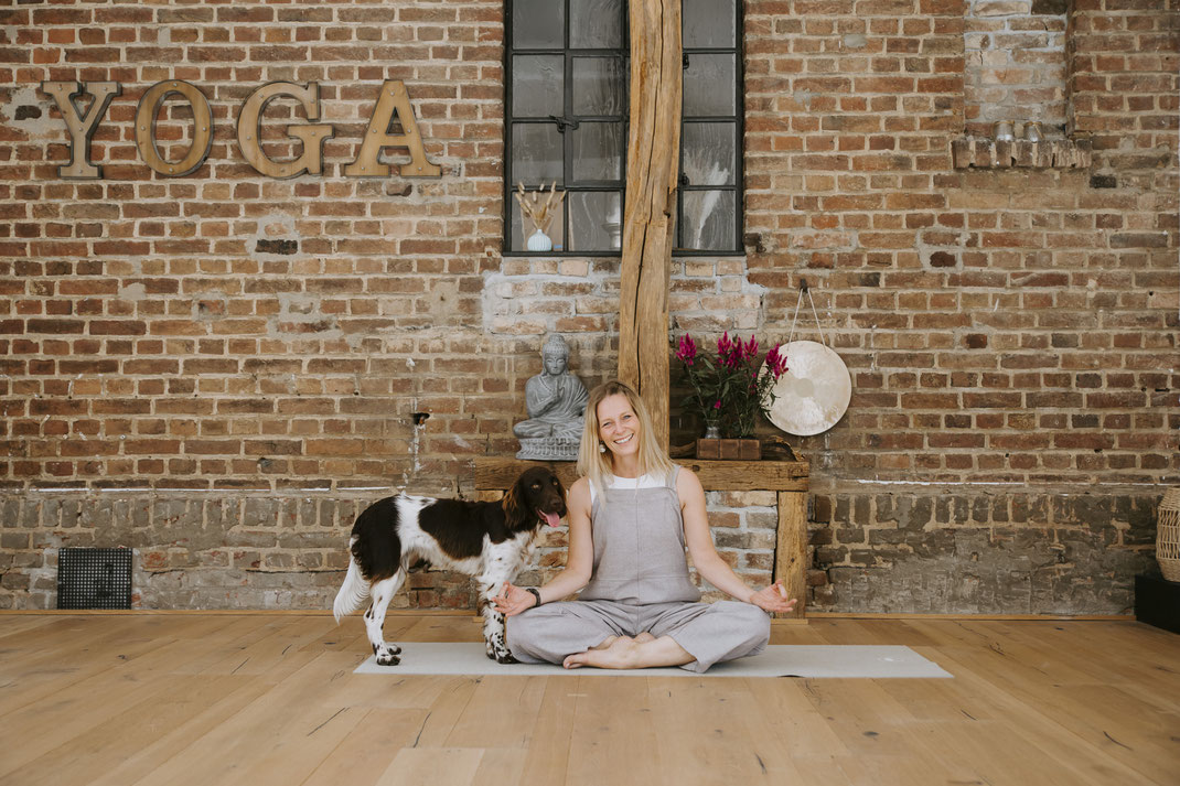 Lachende Frau im Lotussitz im Yogastudio mit Hund an der Seite