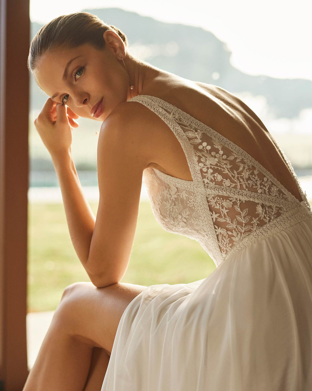 Junge elegante Frau sitzt vor einem großen Fenster in die Kamera blickend und trägt das Brautkleid Zito aus der Aire Boho Kollektion 2026