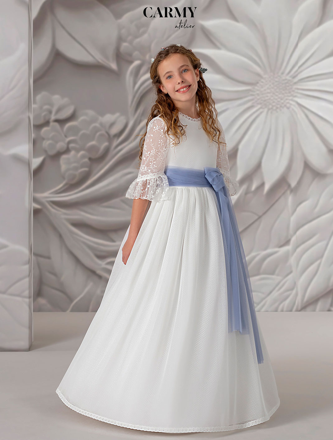 Lächelndes Mädchen trägt ein Kommunionkleid mit einer blauen Schleife aus der Kollektion Carmy Kommunion 2025