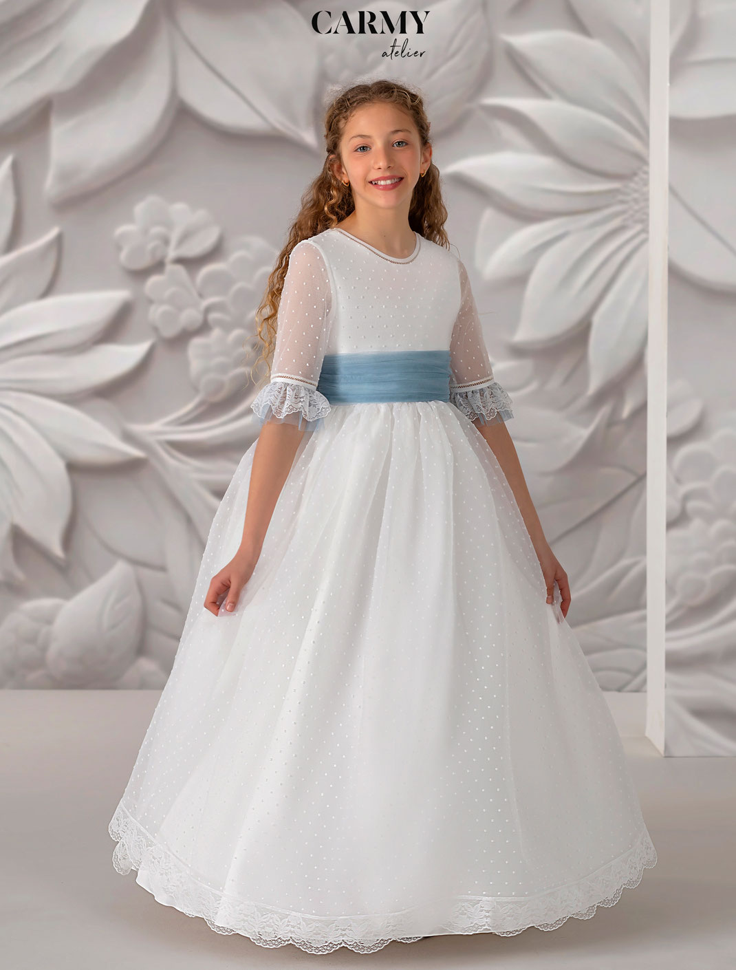 Mädchen trägt ein Kommunionkleid mit blauem Hüftband aus der Carmy Kommunion 2025 Kollektion