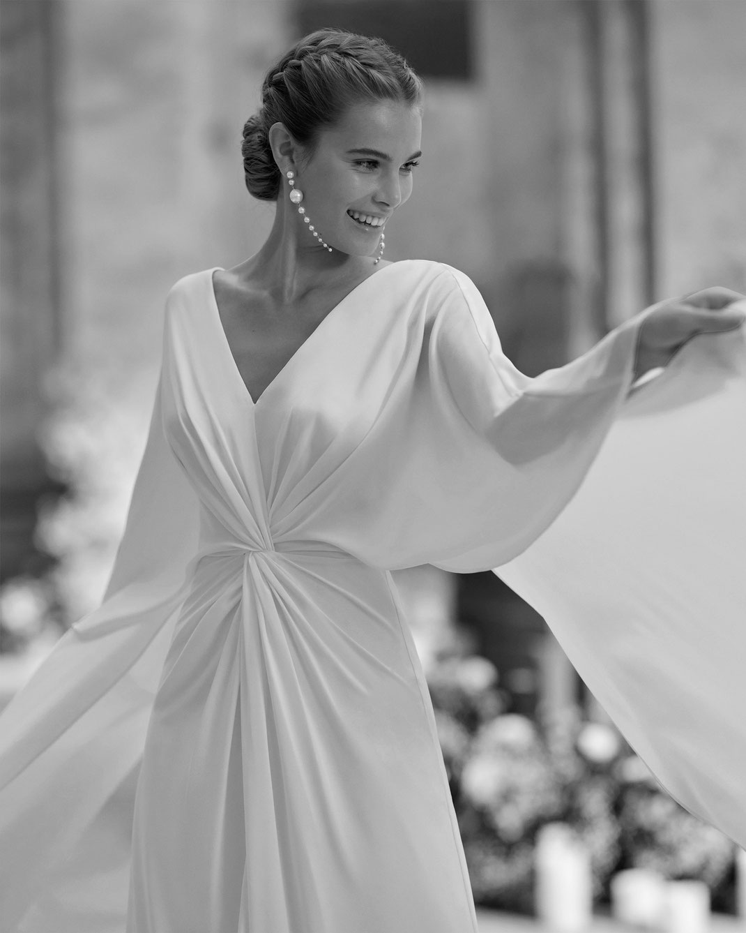 Junge Frau vor Blumen trägt das Brautkleid Halaia aus der Alma Novia Kollektion 2025