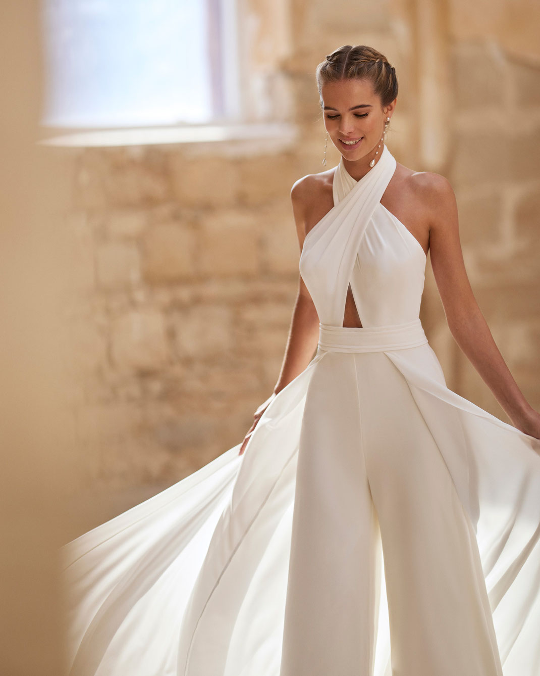 Junge Frau mit Flechtfrisur trägt das Jumpsuit Brautkleid Hadara aus Alma Novia Kollektion 2025