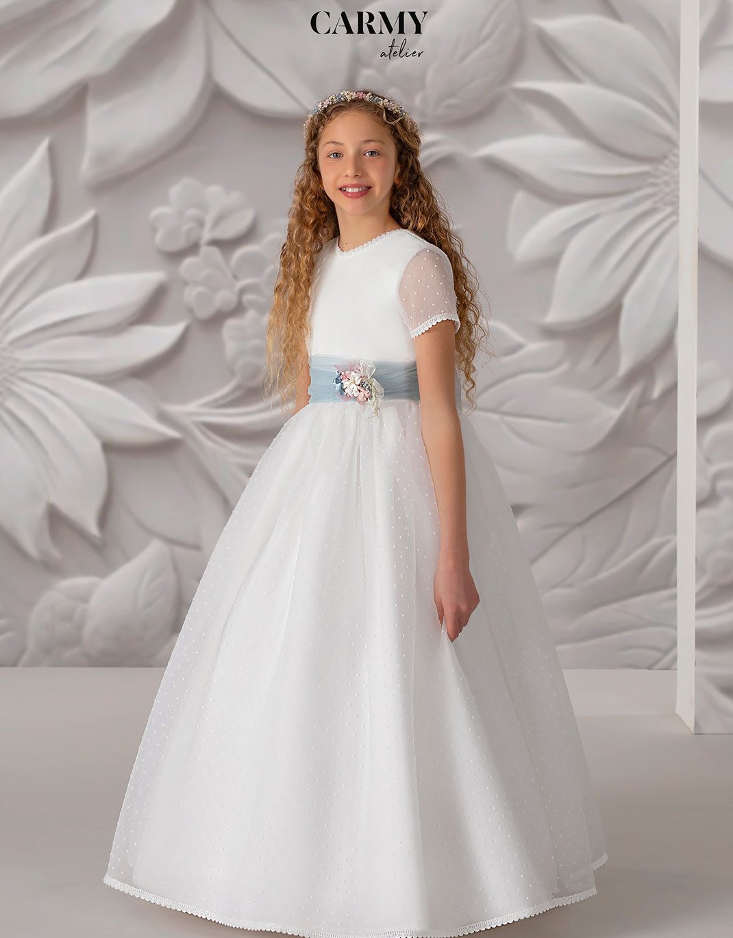 Hübsches Mädchen trägt ein Kommunionkleid mit Blumenschmuck im Haar aus der Carmy Kommunion 2025 Kollektion