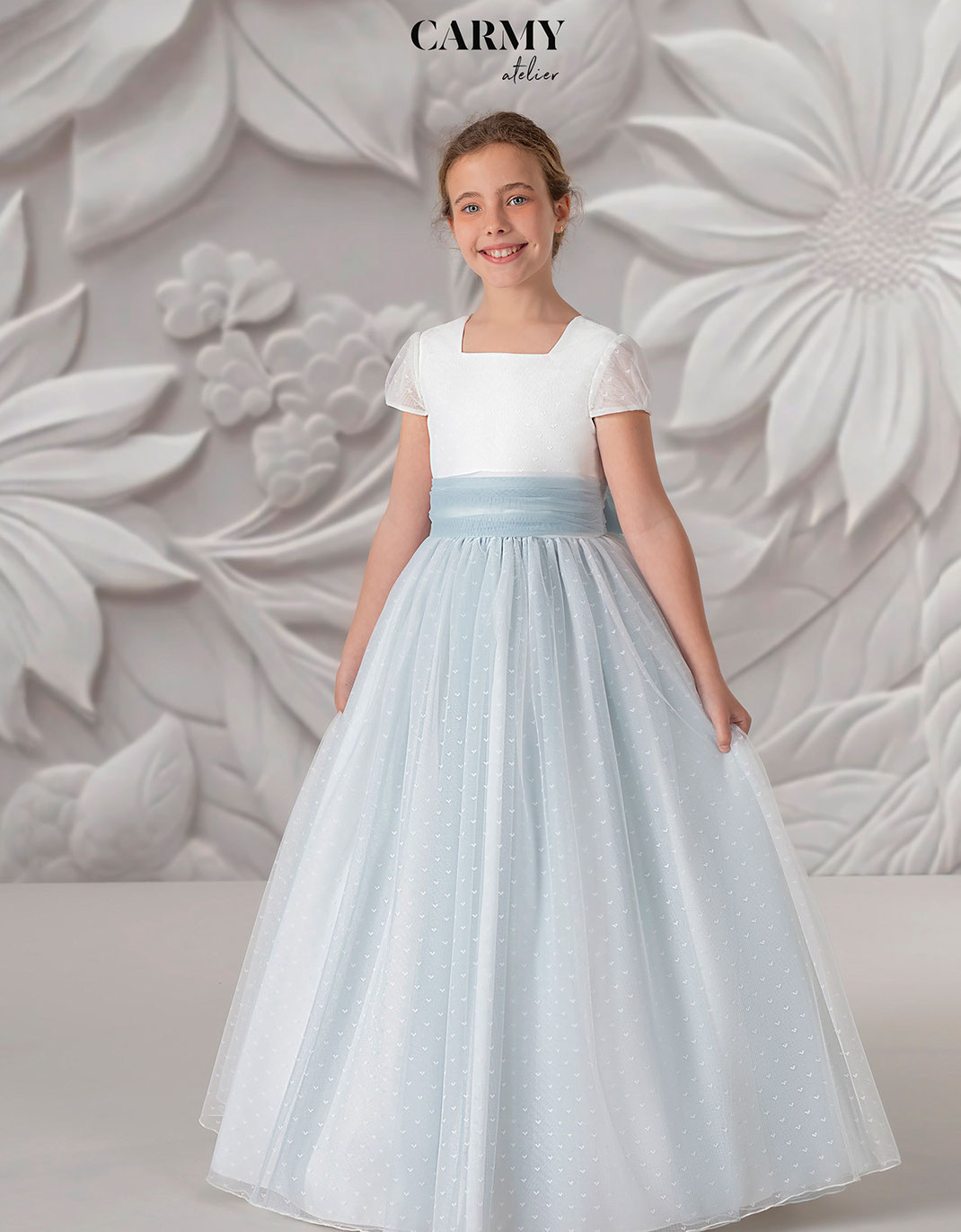 Mädchen trägt ein Kommunionkleid mit blauer Schleife aus der Carmy Kommunion 2025 Kollektion