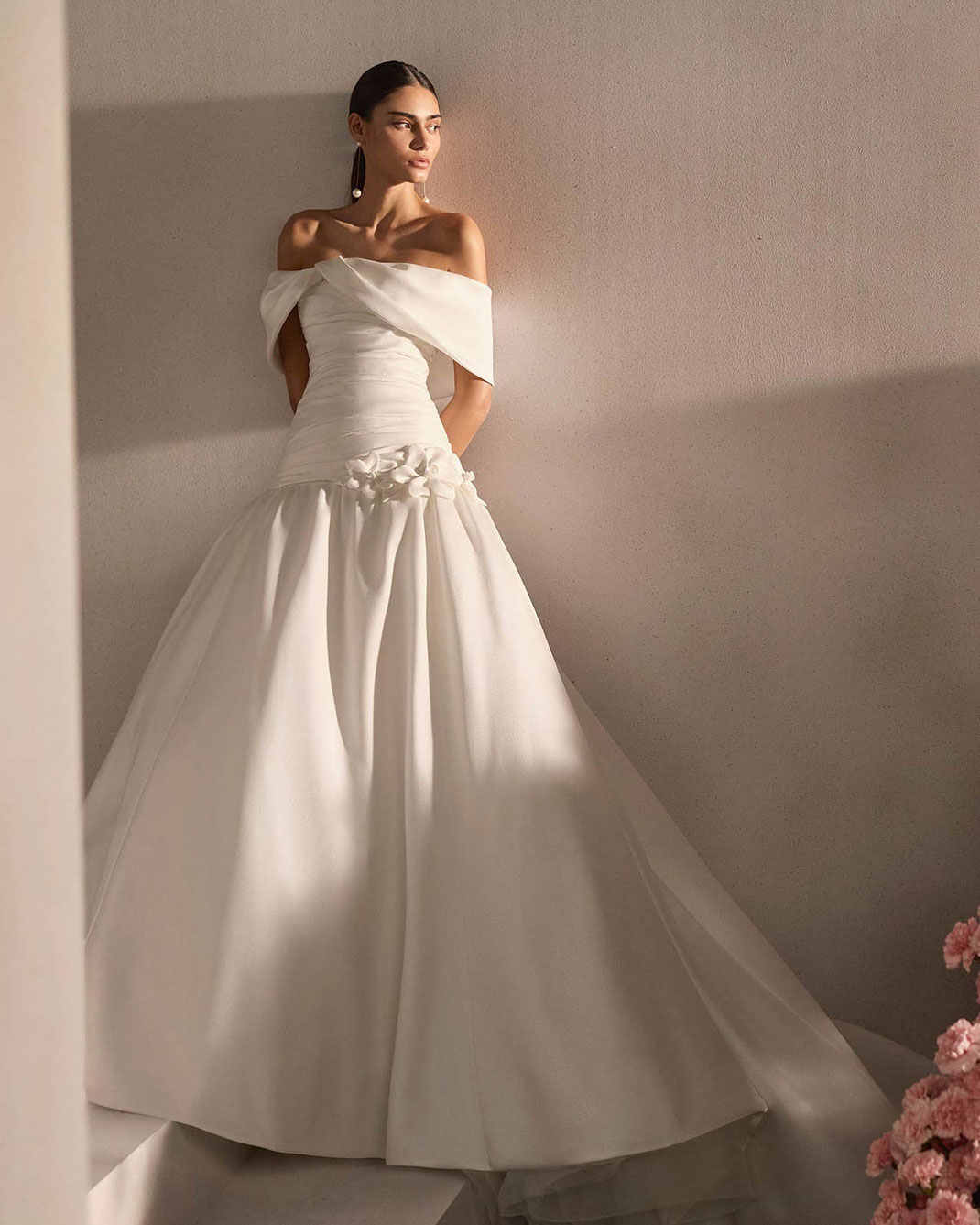 Junge elegante Frau neben rosa Blumen trägt das Brautkleid Rayma von Rosa Clará Couture
