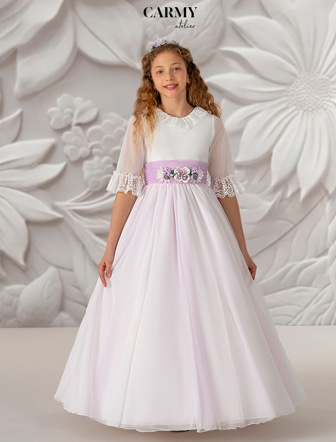 Mädchen mit lockigen Haar trägt ein Kommunionkleid mit Rosa Bund und Blumenschmuck von Carmy aus der Kommunion Kollektion 2025