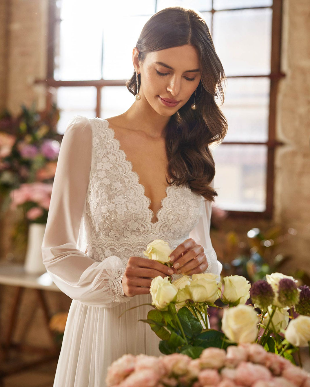 Junge Frau vor Blumen trägt das Brautkleid Xemira aus der Rosa Clará Boheme Kollektion 2026