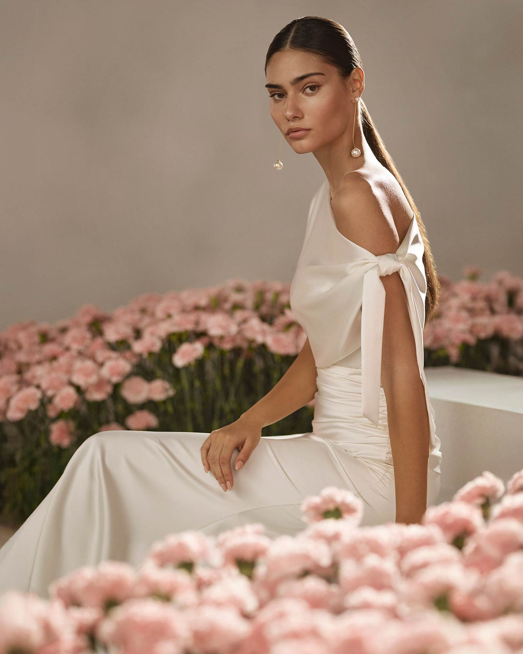 Junge schöne Frau sitzt in einem rosa Blumenmeer und trägt das Brautkleid Rodas aus der Rosa Clará Couture Kollektion 2026