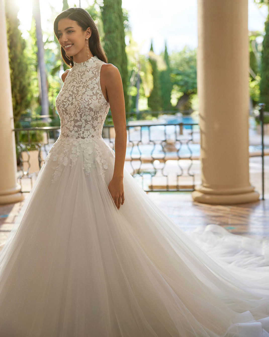 Junge Frau mit braunen Haaren in einem luxuriösen Schlossgarten trägt das Brautkleid Iselda aus der Aire Royale 2025 Kollektion