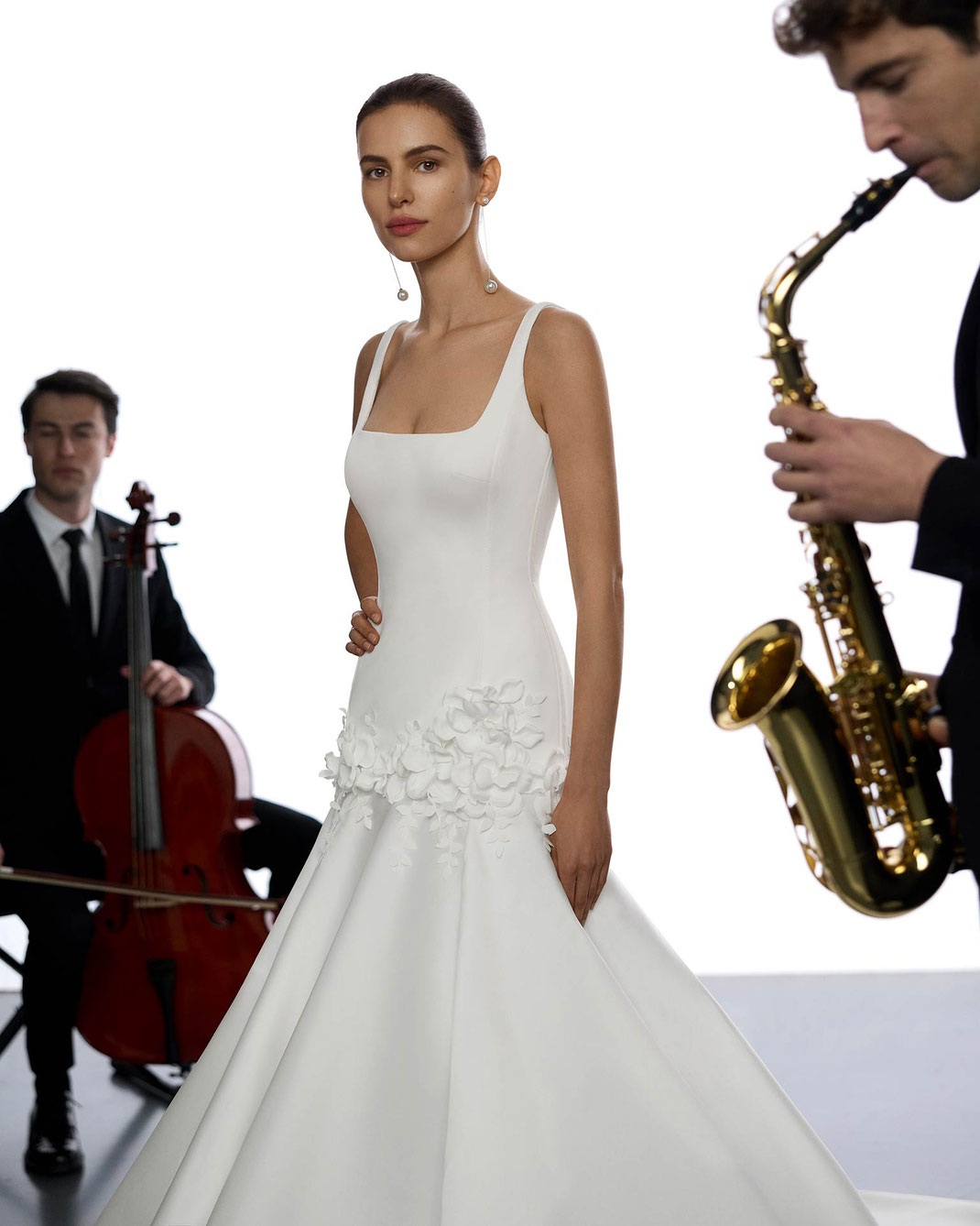 Junge Frau mit glatten braunen Haaren trägt das Brautkleid Yoli aus der Aire Atelier Kollektion 2026 und steht zwischen zwei Musikern