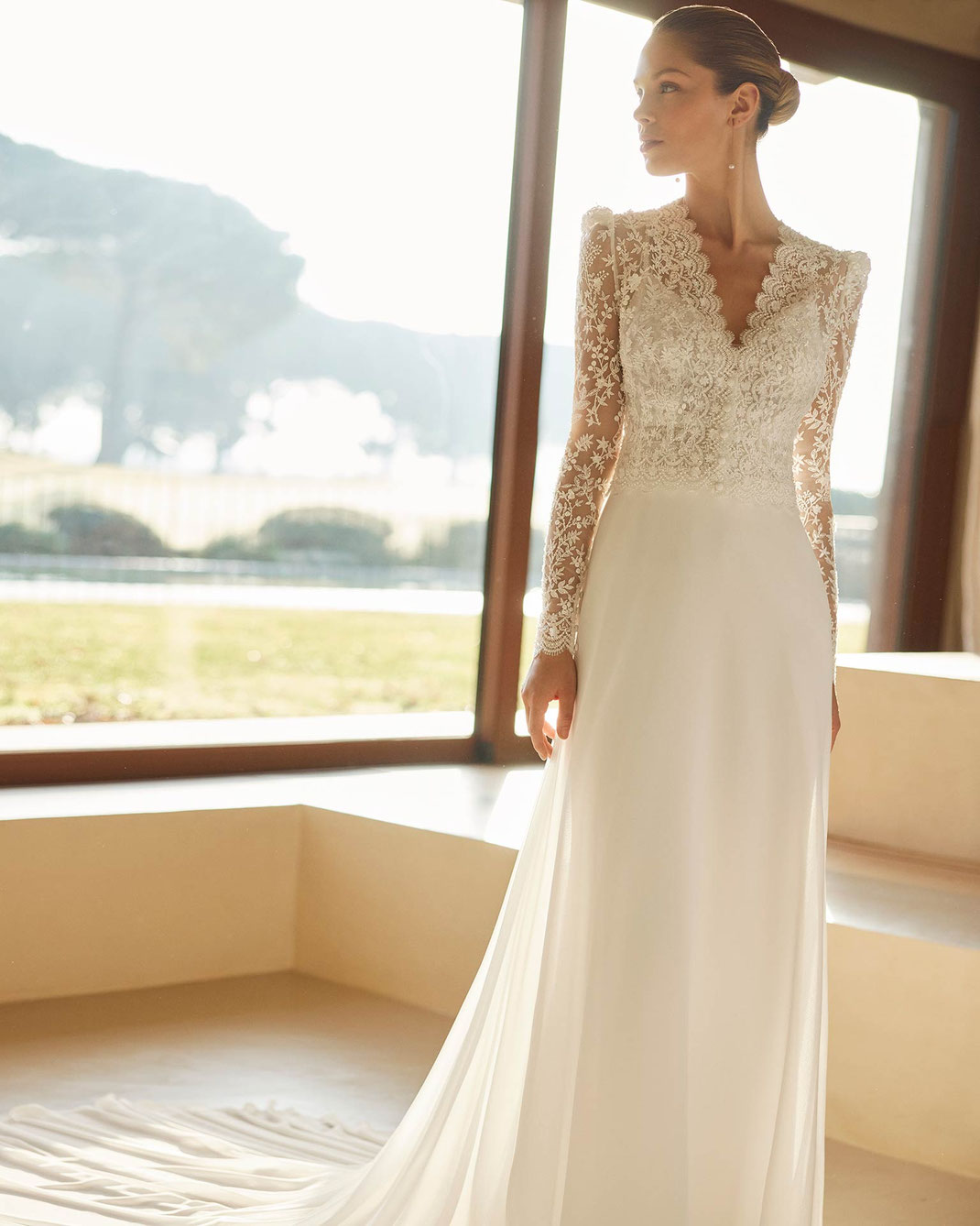 Junge elegante Frau steht vor einem großen Fenster und trägt das Brautkleid Zazurk aus der Aire Boho Kollektion 2026