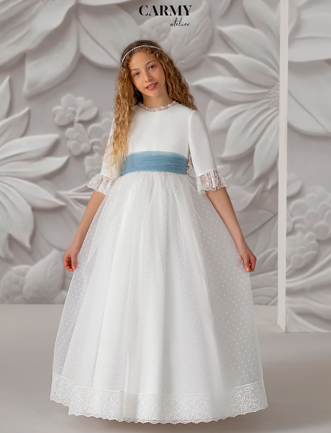 Mädchen trägt ein elegantes Kommunionkleid mit blauer Schleife aus der Carmy Kommunion 2025 Kollektion