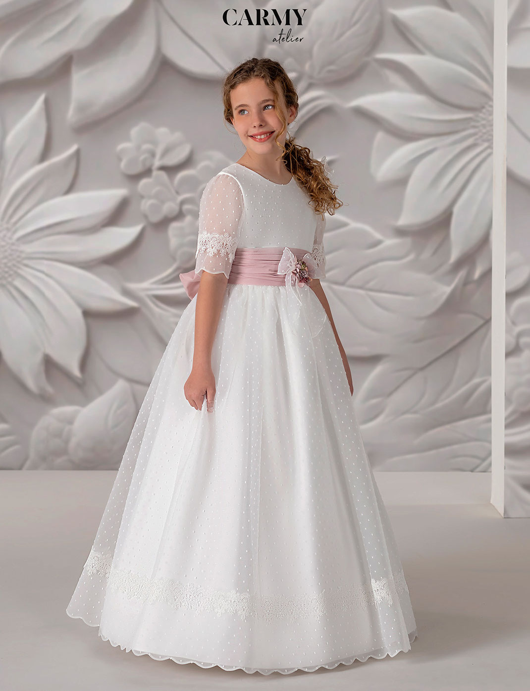 Mädchen trägt ein elegantes Kommunionkleid mit rosa Schleife aus der Carmy Kommunion 2025 Kollektion