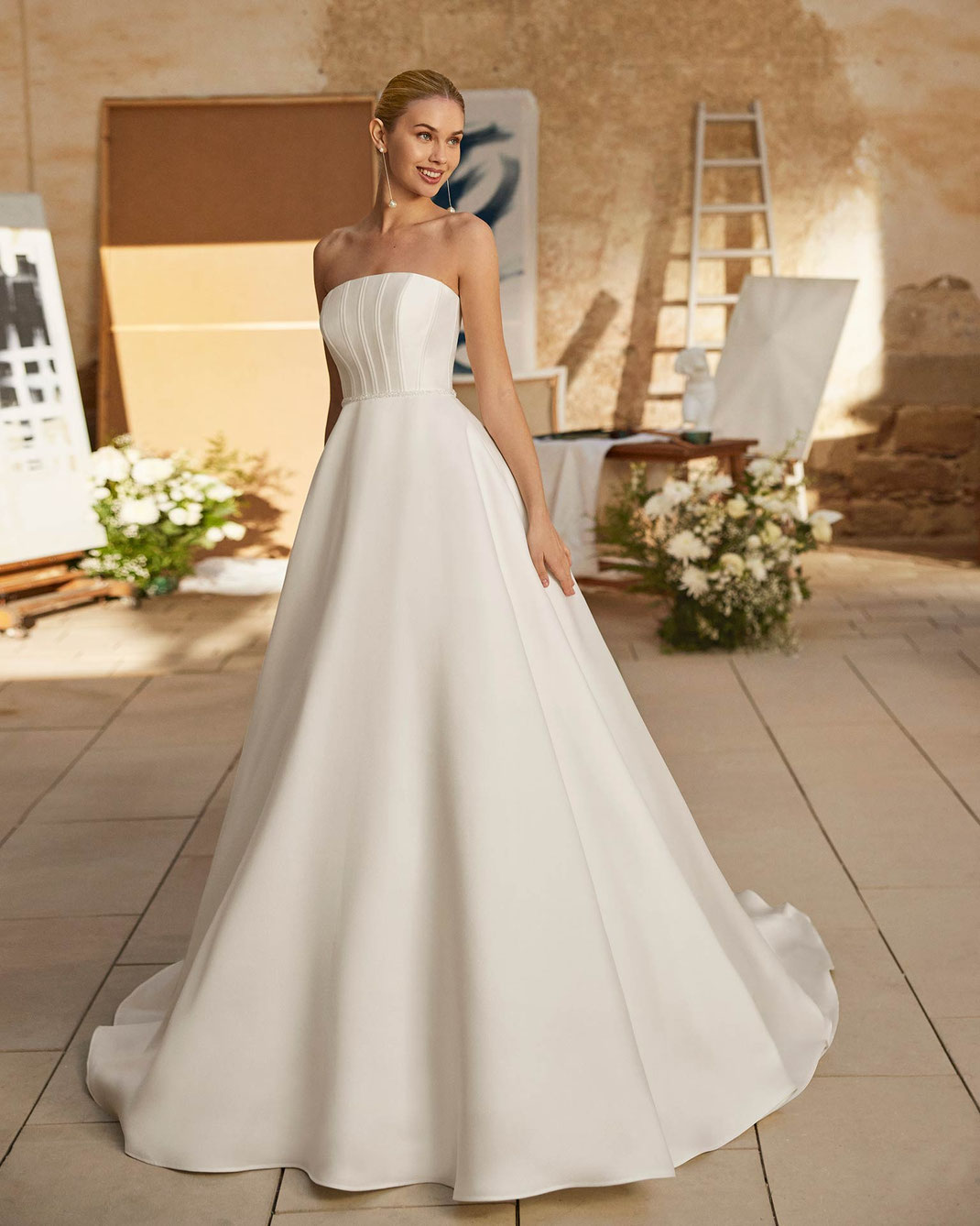 Elegante junge Frau trägt das Brautkleid Moxie in einem Atelier aus der Aire Barcelona Kollektion 2026