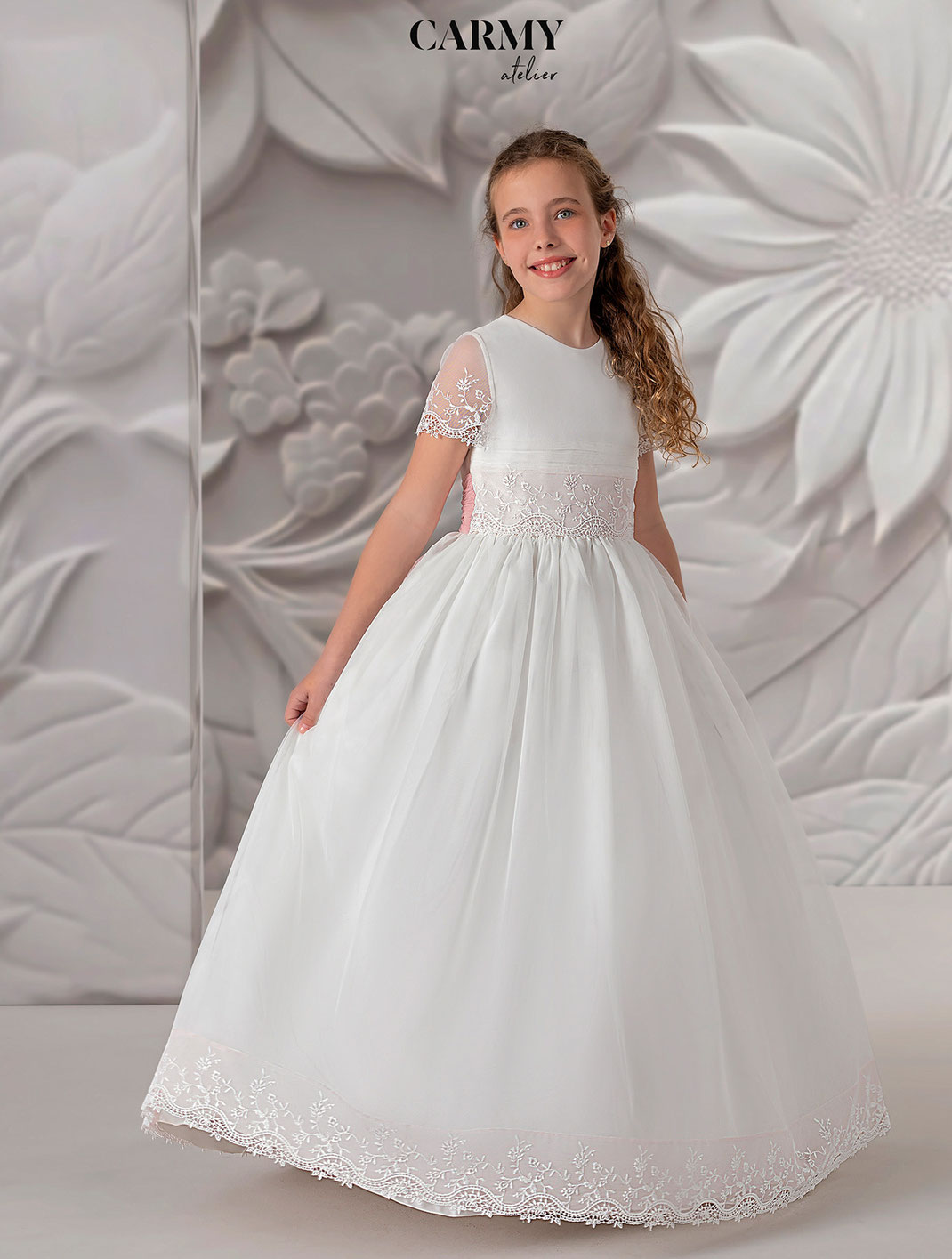 Mädchen mit braunen Haaren trägt ein Kommunionkleid mit rosa Schlaufe aus der Carmy Kommunion Kollektion