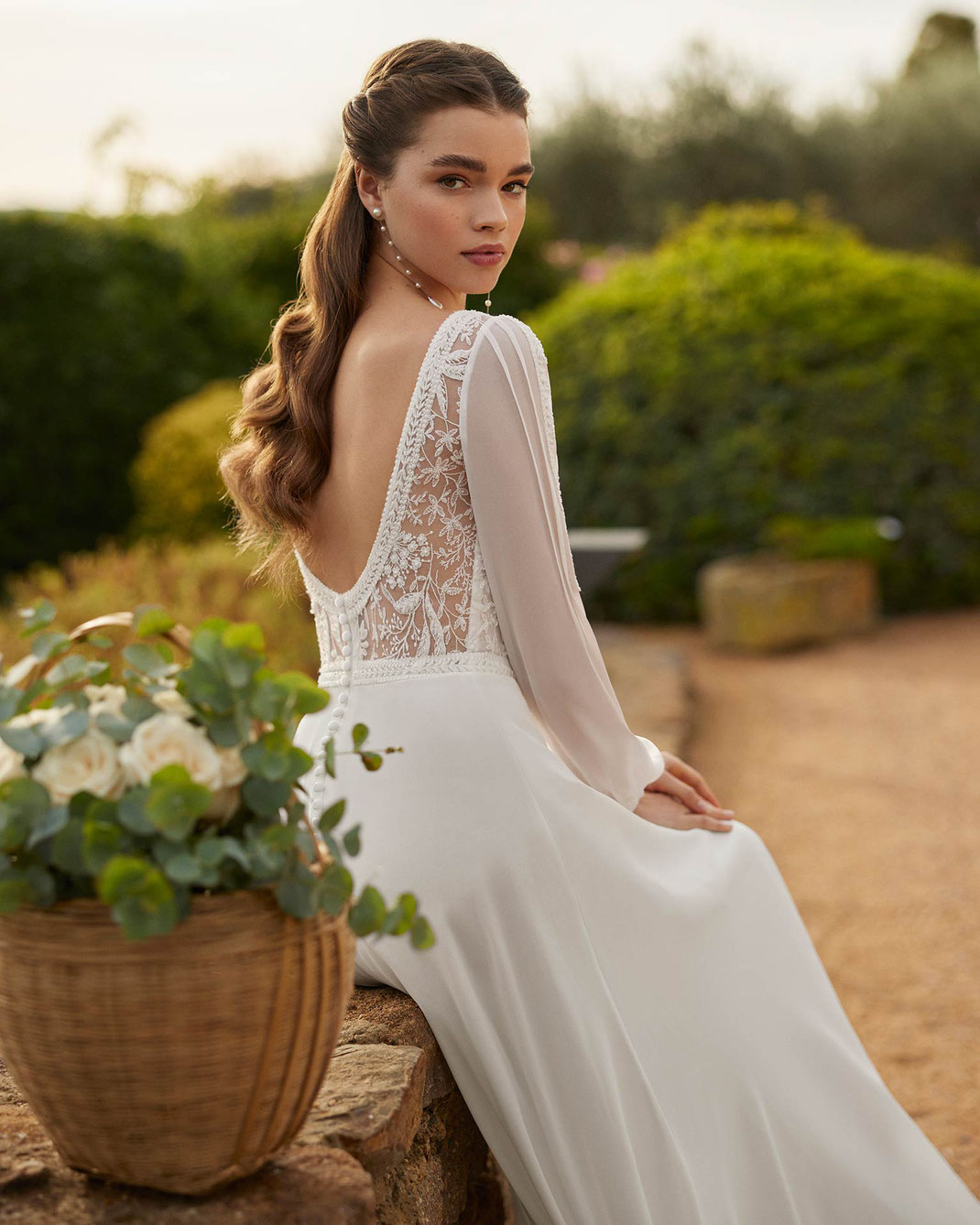 Junge Frau sitzt auf Steinmauer und trägt das Brautkleid Godiva aus der Rosa Clará Boheme Kollektion 2025