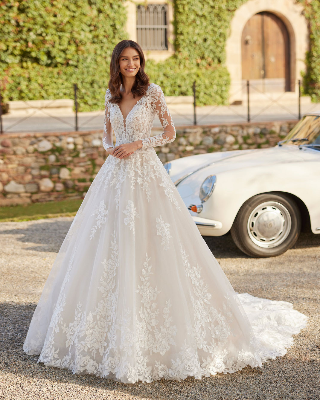 Junge Frau vor einem Oldtimer trägt das Brautkleid Yaony aus der Aire Royale Kollektion