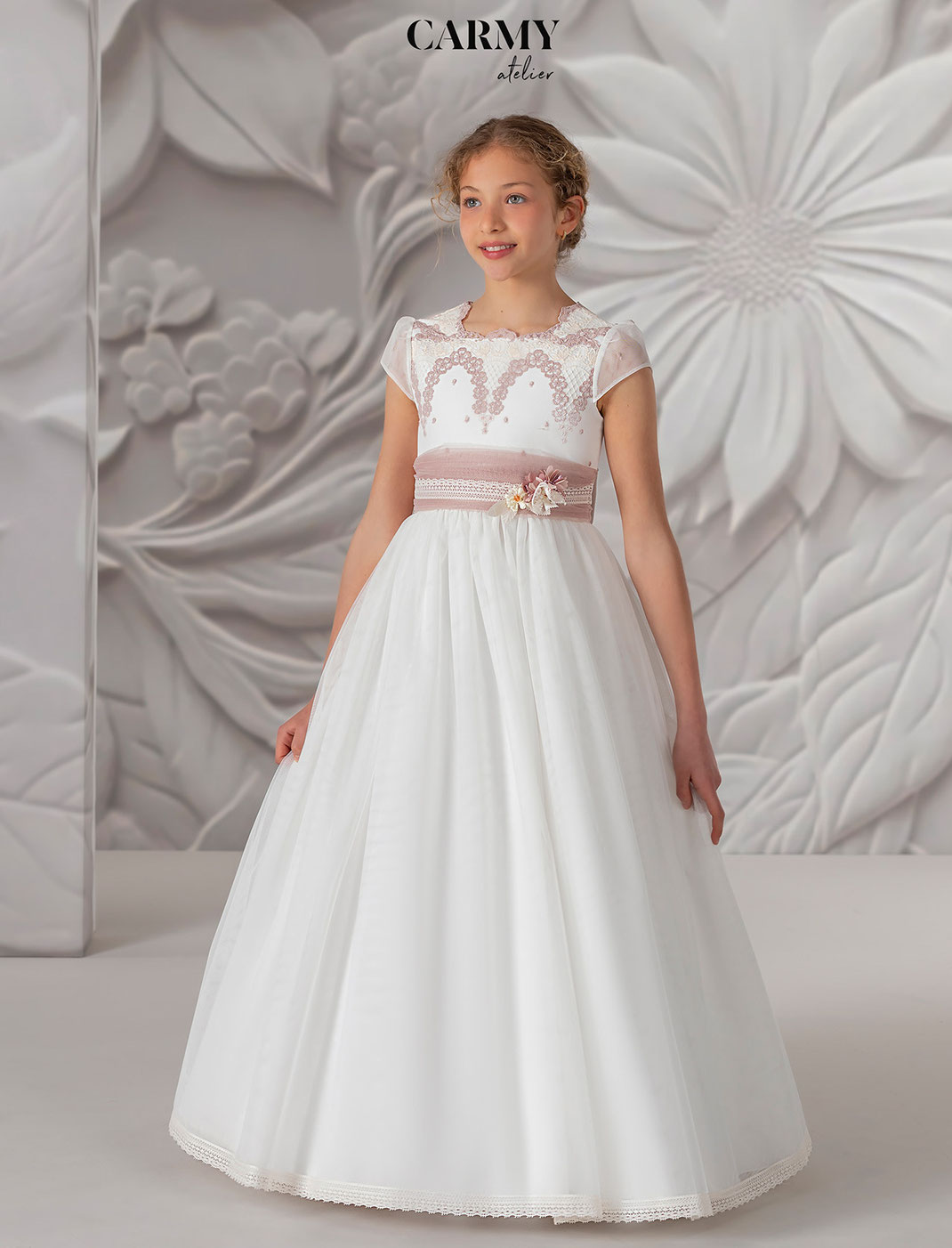 Lächelndes Mädchen trägt ein Carmy Kommunionkleid Kollektion verziert mit rosa Elementen