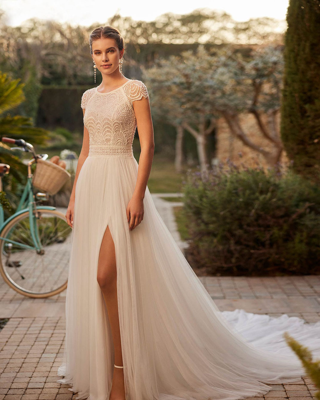 Junge Frau vor Fahrrad trägt ein elegantes, ausgestelltes Brautkleid aus weichem Tüll, Spitze und Strass von Rosa Clara