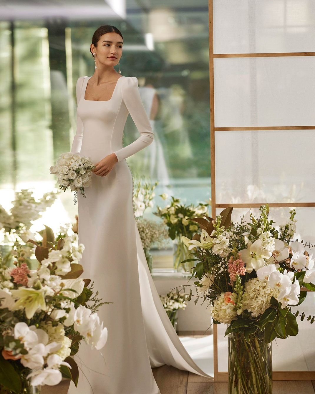 Junge Frau mit braunen Haaren zwischen Blumen trägt das Brautkleid Marvin aus der Rosa Clará Couture Kollektion 2026