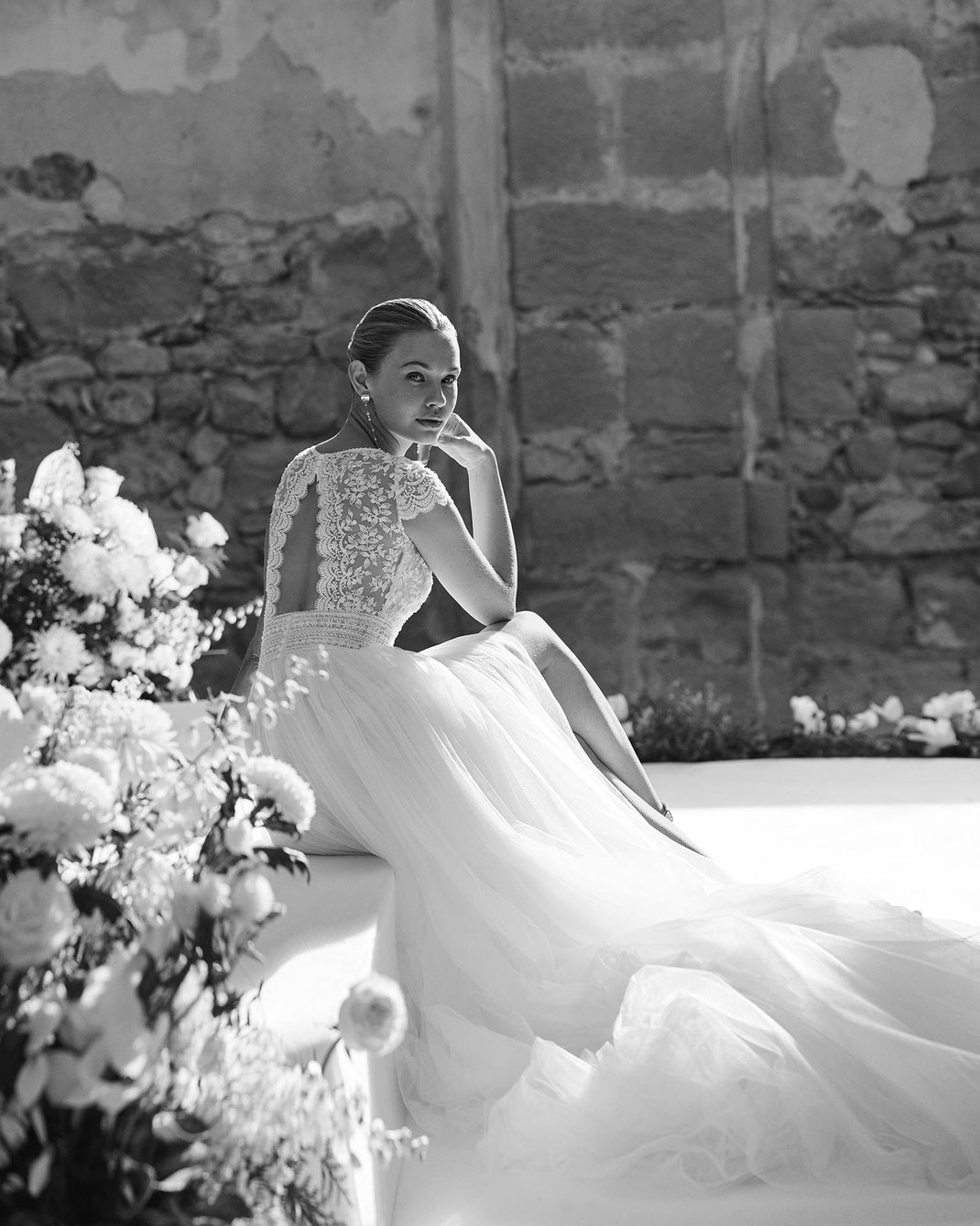 Elegante Frau hinter Blumen sitzend trägt das Brautkleid Melina aus der Aire Barcelona Kollektion 2026