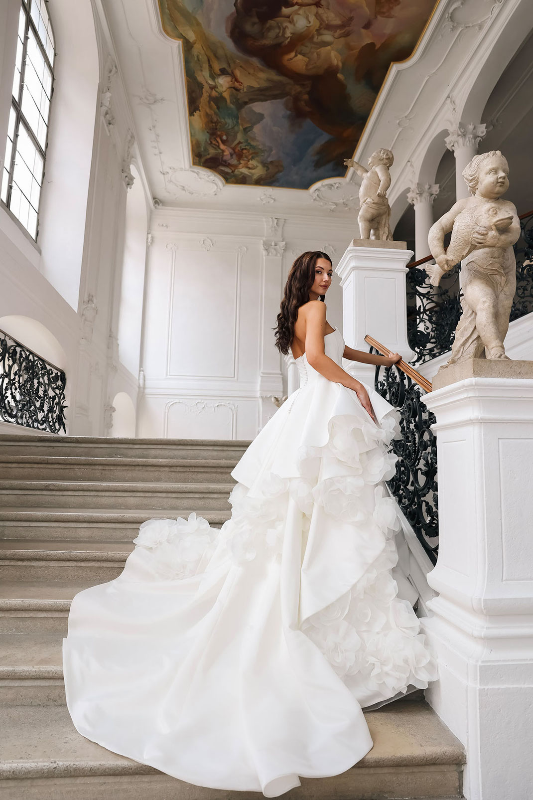 Junge schöne Frau stehend auf einer klassischen Treppe in einer Villa trägt ein Brautkleid von Amo & Luv