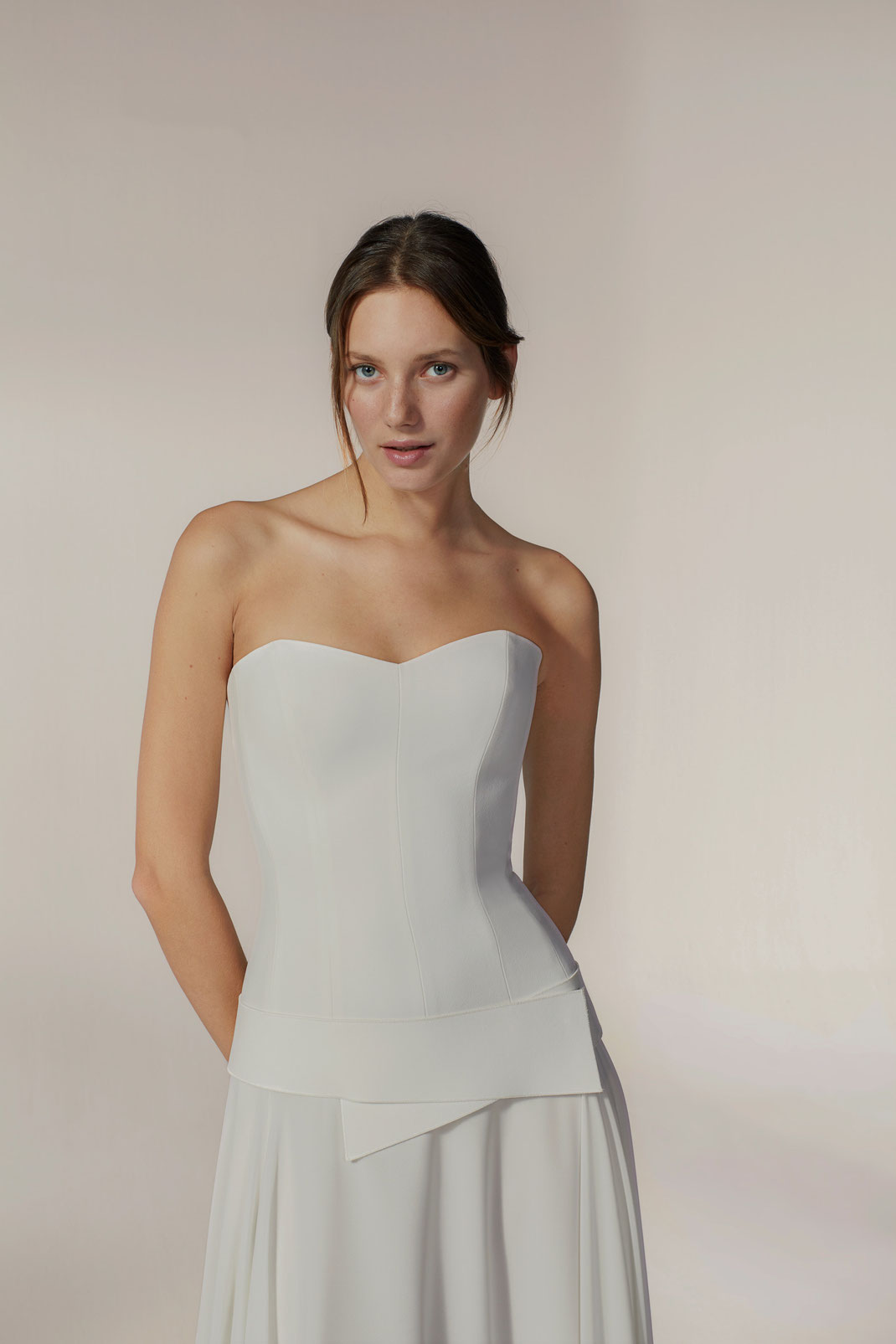 Junge Frau mit braunen Haaren trägt ein schulterfreies Brautkleid mit tiefer Taille, Gürtel und Chiffonrock von Jesus Peiro