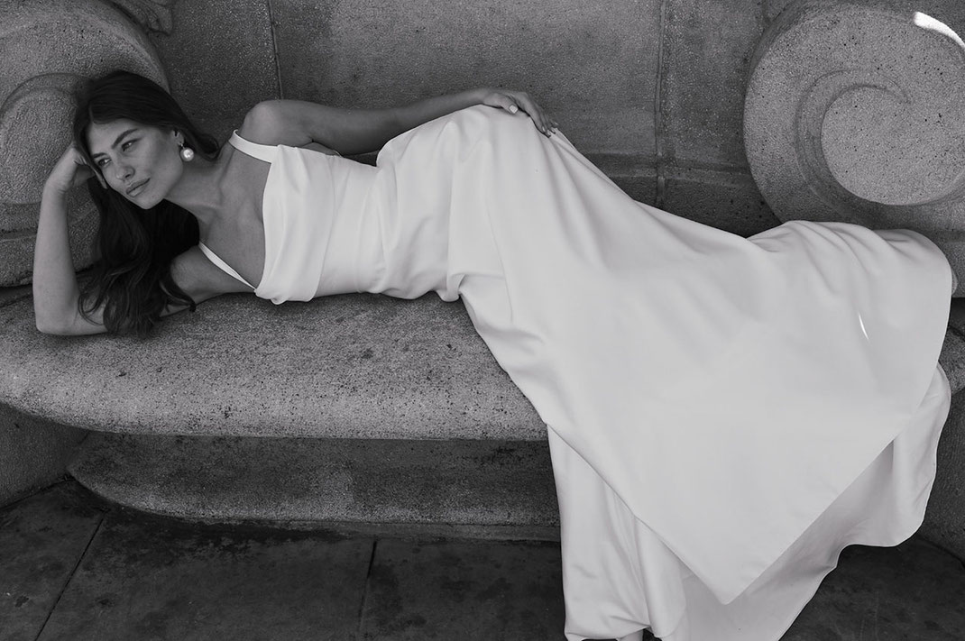 Eine junge Frau liegt mit einem Brautkleid von Jesus Peiro auf einer Parkbank aus Stein