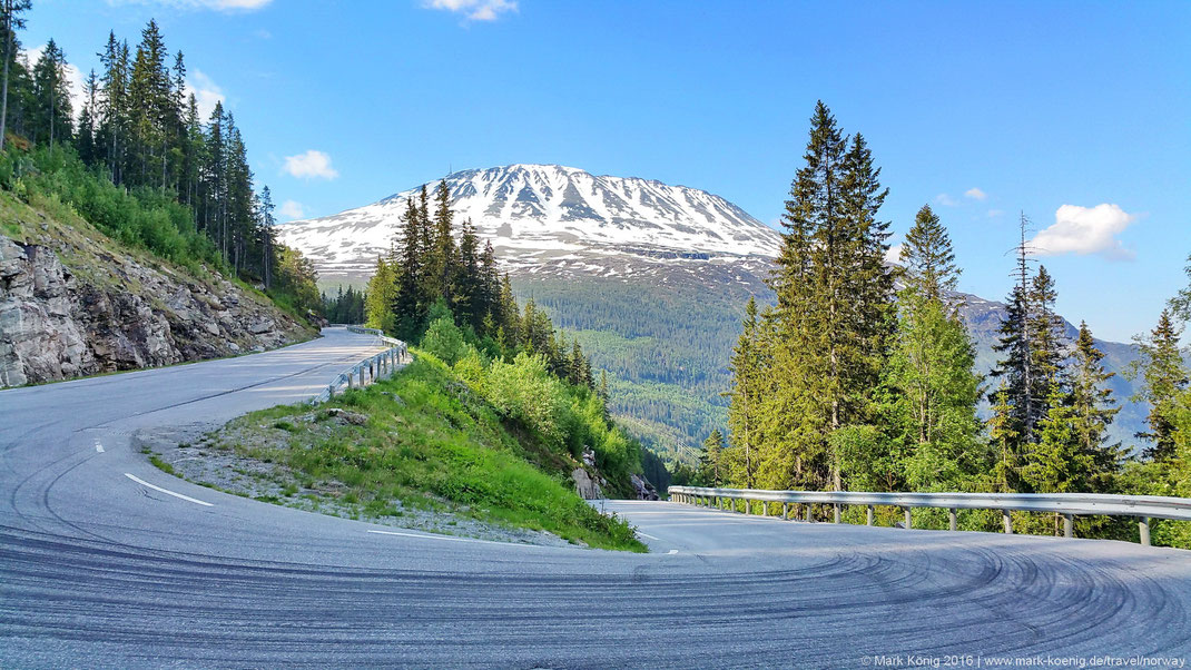 Photo showing snow-capped peak of Gaustatoppen mountain with blue sky; a narrow hairpin curve in the front of the picture