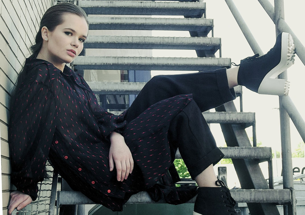 Braunhaarige Frau trägt eine schwarze Jacke mit Ledertaschen und Kapuze. Mund und Nase sind verdeckt