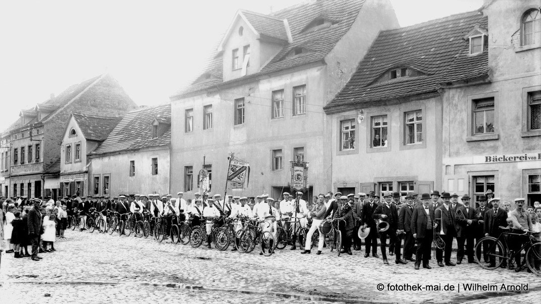 Kleindstadthäuser, auf der Straße Musiker, Radfahrer mit Fahnen, Wappen und Zuschauer. 