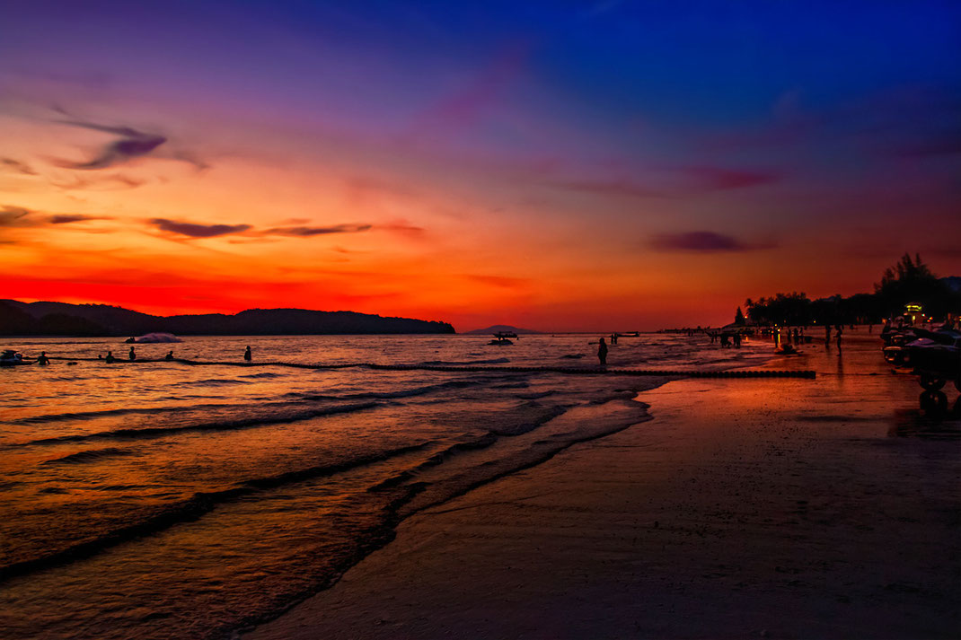 Sonnenuntergang mit orange rotem Himmel am Cenag Beach auf Langkawi-Malaysia. Einige Leute stehen im Wasser oder schwimmen. Fotos aus Malaysia kostenlos downloaden bei www.mjpics.de