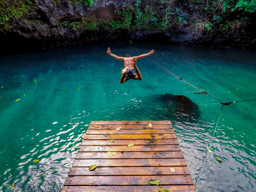 Man jumping into a blue water hole from a wooden pier in a tropical location.