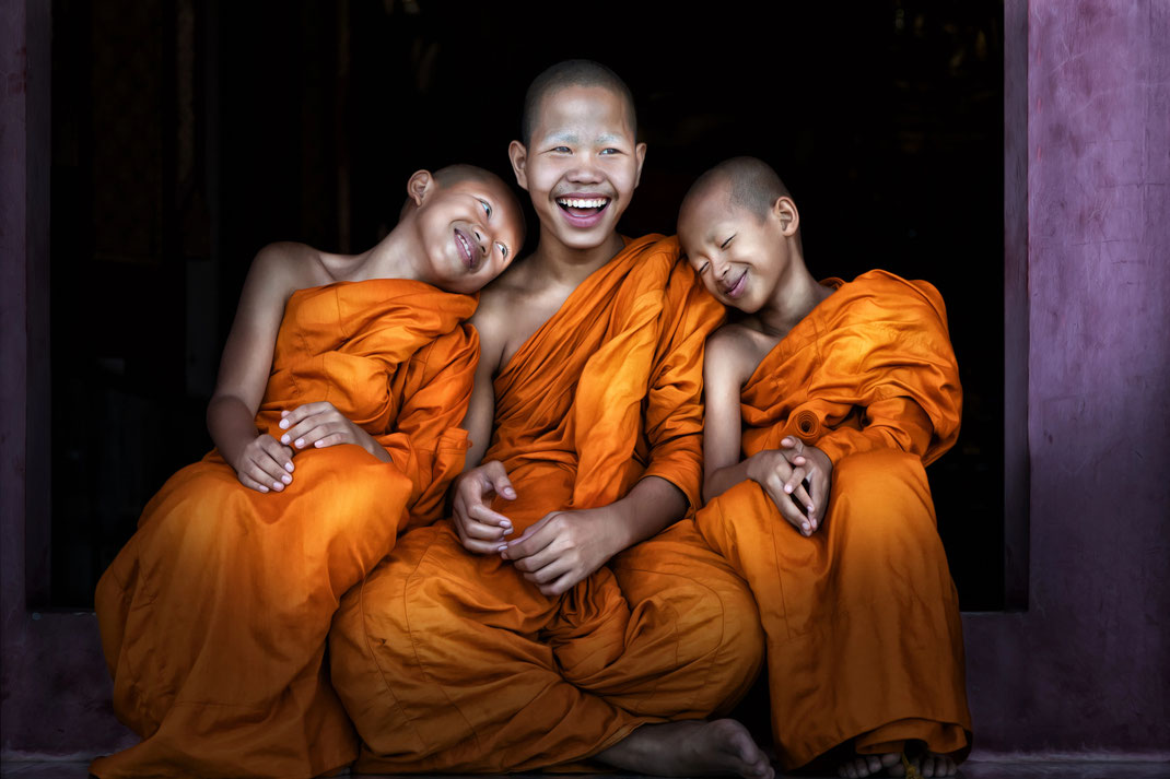 Drei junge, lachende buddhistische Novizen in safrangelben Roben, die fröhlich beieinandersitzen.