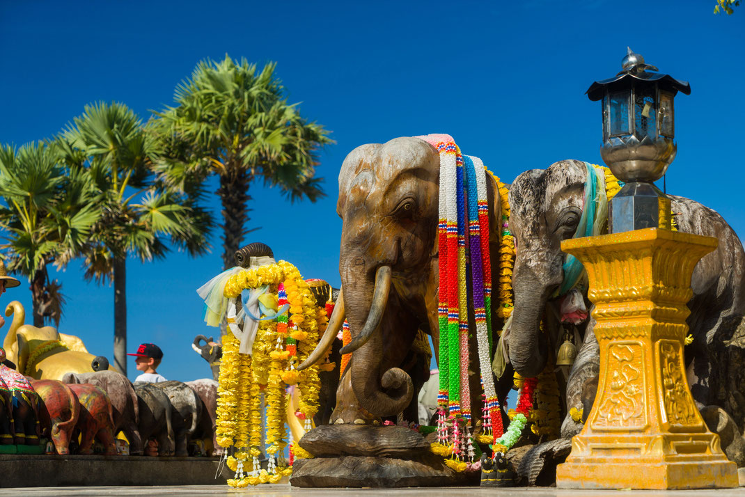 Farbenfroh geschmückte Elefantenstatuen mit leuchtenden Blumengirlanden an einem thailändischen Schrein unter strahlend blauem Himmel.