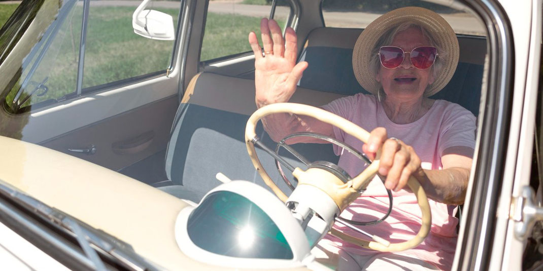 Ältere Frau mit Sonnenbrille und Hut winkt lachend aus einem Oldtimer heraus