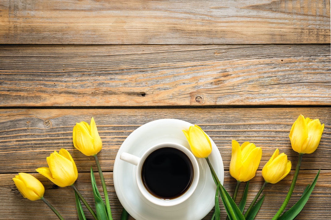 Tasse schwarzer Kaffee auf weißer Untertasse, umgeben von gelben Tulpen auf rustikalem Holztisch – Frühlingshafte Frühstücksszene in der Draufsicht