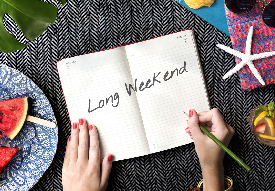 Aufgeschlagenes Notizbuch mit der Aufschrift „Long Weekend“, daneben sommerliche Accessoires wie Wassermelone, Cocktail und Seestern – Urlaubsplanung in entspannter Atmosphäre.