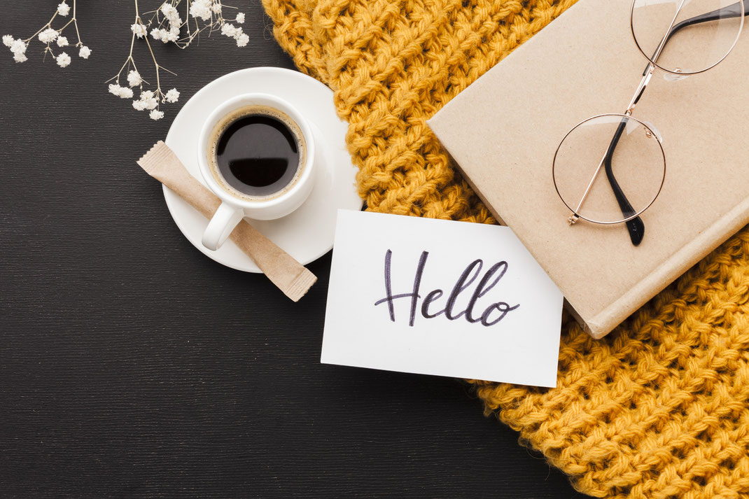 Stimmungsvolle Morgenroutine mit schwarzem Kaffee, Notizkarte mit Hello-Schriftzug, Notizbuch und Brille auf gelbem Strick – gemütlicher Start in den Tag