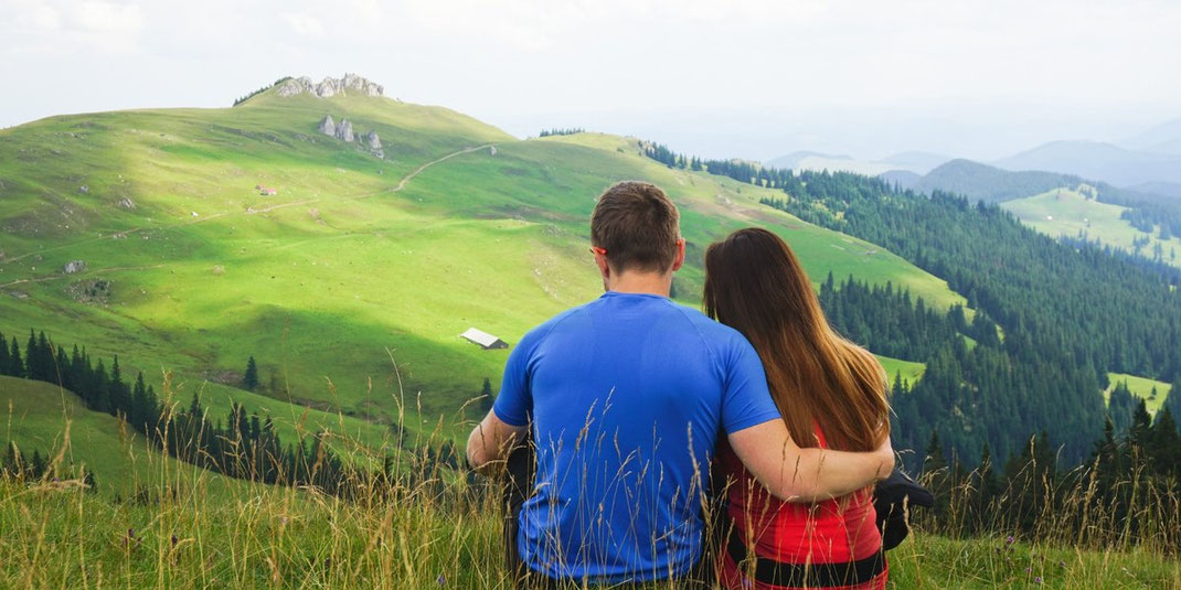 Paar sitzt eng umschlungen auf einer Bergwiese und schaut gemeinsam ins Tal