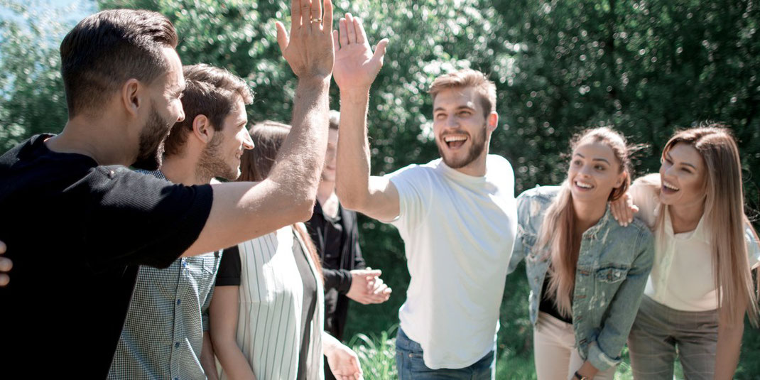 Gruppe junger Menschen im Freien, zwei Männer geben sich ein High-Five, andere lachen und schauen zu