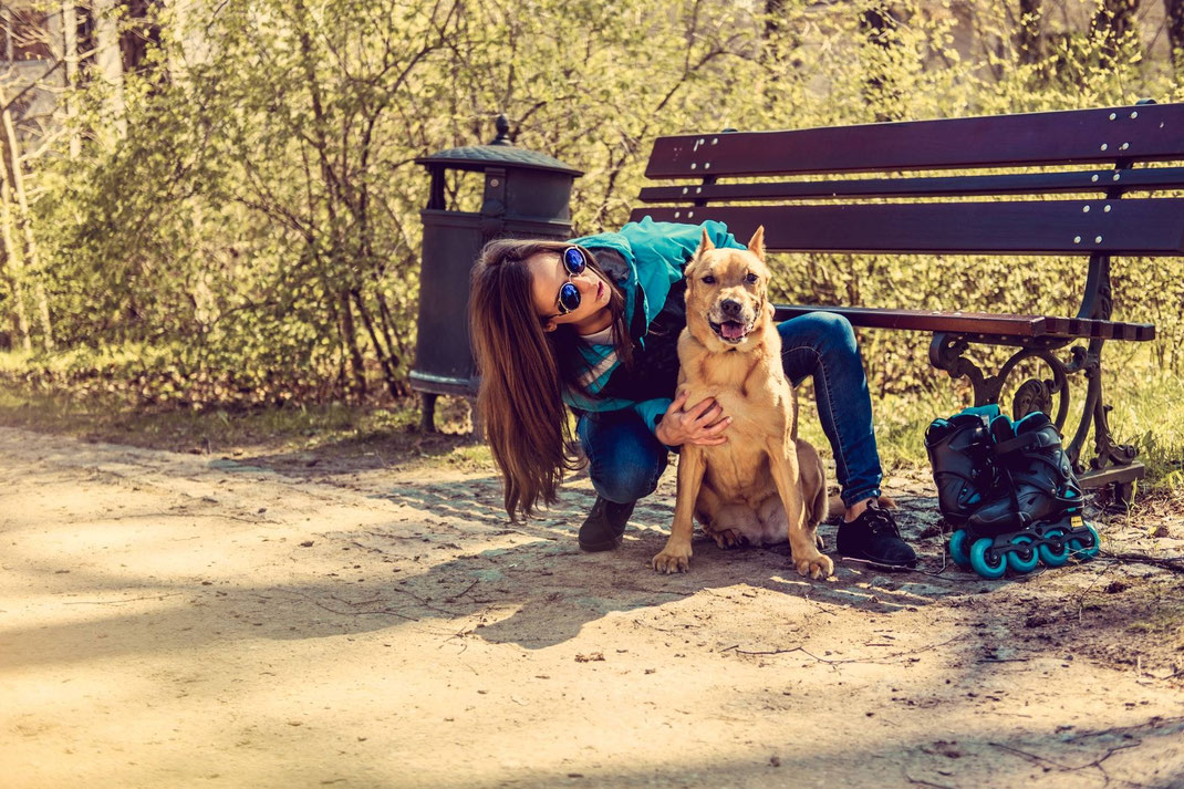 Frau mit Sonnenbrille umarmt ihren Hund auf einem Parkweg neben einer Bank und Inlineskates.