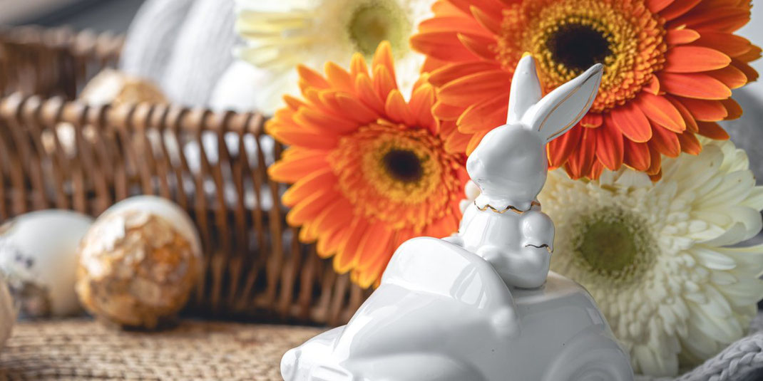 Weiße Keramik-Osterhasen mit bunten Eiern und lila Blüten auf Leinenstoff.