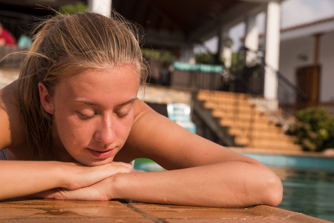 Junge Frau liegt entspannt am Rand eines Pools und genießt die Sonne mit geschlossenen Augen.